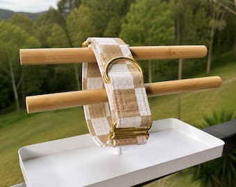 Puede incluir: Un collar para mascota con un patrón de cuadros beige y blanco hecho de tela. El collar incluye una anilla en D de metal dorado y una hebilla deslizante ajustable a juego de metal dorado, exhibido en un soporte de madera con bandeja blanca.