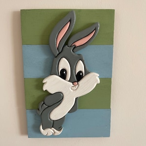 May include: A wooden wall art piece featuring a cartoon rabbit with gray fur, white fur on its chest, and pink ears. The rabbit is painted on a multi-colored background with stripes of green and blue.