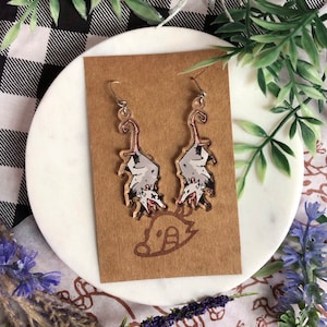 May include: A pair of dangle earrings featuring a cartoon illustration of a possum. The possum is white with black markings and has a pink nose. The earrings are made of clear acrylic and have silver hooks.