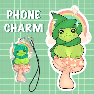 May include: A green frog wearing a green hat and scarf sits on a pink mushroom with white hearts. The frog is a phone charm with a rainbow and glitter background.