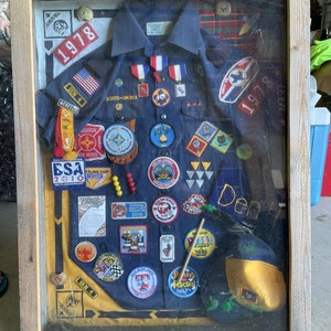 Puede incluir: Una vitrina enmarcada de un uniforme azul de Boy Scout con varios parches y distintivos. El uniforme tiene un pañuelo de bolsillo amarillo y un sombrero amarillo. El marco es de madera y tiene una placa con el texto "Erik Carman Pack 1978 - ADL 2011".