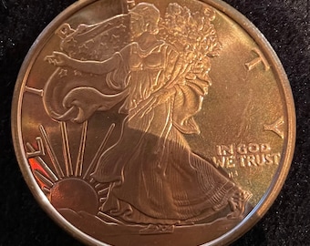 Op de afbeelding: Een bronzen munt met een gedetailleerd reliëf van een vrouw die naar links loopt, een tak vasthoudt, met een opkomende zon op de achtergrond. De munt heeft de tekst "LIBERTY" en "IN GOD WE TRUST".