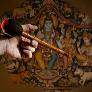 Op de afbeelding: Een houten percussie-instrument met een donkerbruine kop en een lange, gepolijste steel. Het instrument wordt vastgehouden voor een kleurrijke muurschildering met religieuze figuren en dieren.
