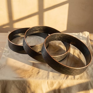 May include: Three abstract decorative rings with a dark wood grain finish and copper-colored edges. The rings are arranged in a flowing, overlapping design, creating a modern and artistic display. The rings are displayed on a neutral-colored surface.