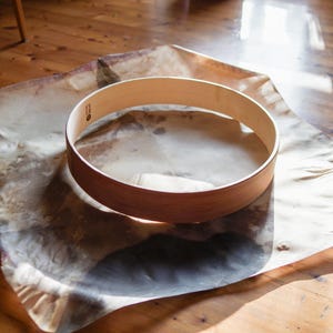 Peut inclure: Un cercle de tambour en bois brun clair repose sur une peau d'animal. Le cercle de tambour est circulaire et a une finition lisse et polie. La peau est brun clair et beige avec un motif moucheté. Le fond est un plancher en bois.