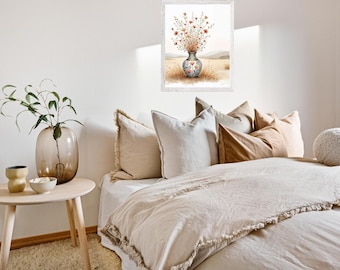 May include: A framed watercolor print of a vase filled with wildflowers. The vase has a blue and white floral design. The print hangs above a bed with beige bedding and pillows. A glass vase with greenery and a small wooden side table are in the foreground.