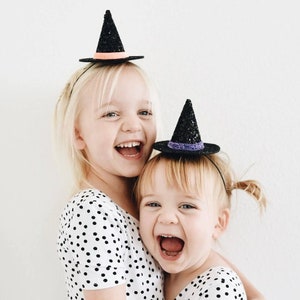 May include: Two young girls wearing black witch hats with glitter. The girl in the front is wearing a white polka dot shirt and the girl in the back is wearing a white polka dot dress. Both girls are smiling.