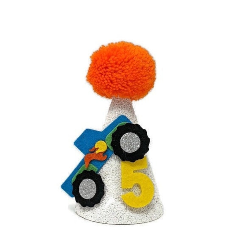 Op de afbeelding: Een witte feestmuts met een oranje pompon bovenop. De muts heeft een blauw monstertruck-ontwerp met het nummer 5 op de voorkant.