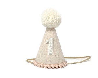 Peut inclure: Un chapeau de fête en feutre rose avec un pompon blanc sur le dessus et un chiffre "1" en feutre blanc sur le devant. Le chapeau est orné d'une bordure de pompons roses autour du bas.