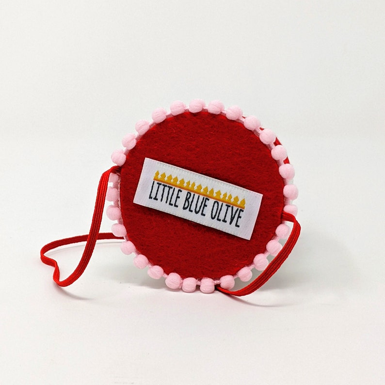 May include: A red felt circle with a pink pom-pom trim and a white label that says "Little Blue Olive". The circle is attached to a red strap.
