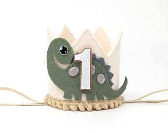 May include: A cream-colored felt crown with a green dinosaur and the number 1. The dinosaur has gray spots and a smiling face. The crown has a tan pom-pom trim and an elastic band.