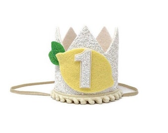 May include: White felt crown with a yellow felt lemon and green felt leaves. The lemon has a silver glitter number 1 on it. The crown has a white tassel trim.