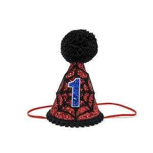 Puede incluir: Sombrero de fiesta rojo y negro con purpurina y un pompón negro en la parte superior. El sombrero tiene un diseño de telaraña y un número uno azul en la parte delantera.