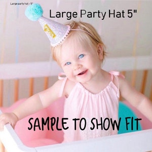 May include: A child wearing a pink dress and a sparkly silver party hat with a blue pom-pom on top. The hat has a gold number one on it. The text "Large Party Hat 5" is visible in the image. The text "SAMPLE TO SHOW FIT" is also visible in the image.
