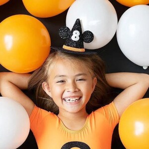 May include: A young girl wearing an orange shirt with a black witch hat with a sparkly number 8 on it. The hat has Mickey Mouse ears. The girl is surrounded by orange and white balloons.