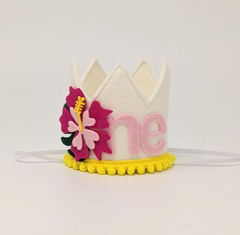 May include: White felt crown with a pink felt "one" and a pink and green felt hibiscus flower. The crown has a yellow pom-pom trim.