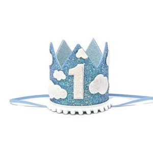May include: Blue felt crown with white felt clouds and a white felt number one. The crown has a white pom-pom trim and a blue elastic band.