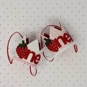 May include: Two white felt crowns with red felt strawberries and the word "one" in red felt letters. The crowns have pink and white pom pom trim.