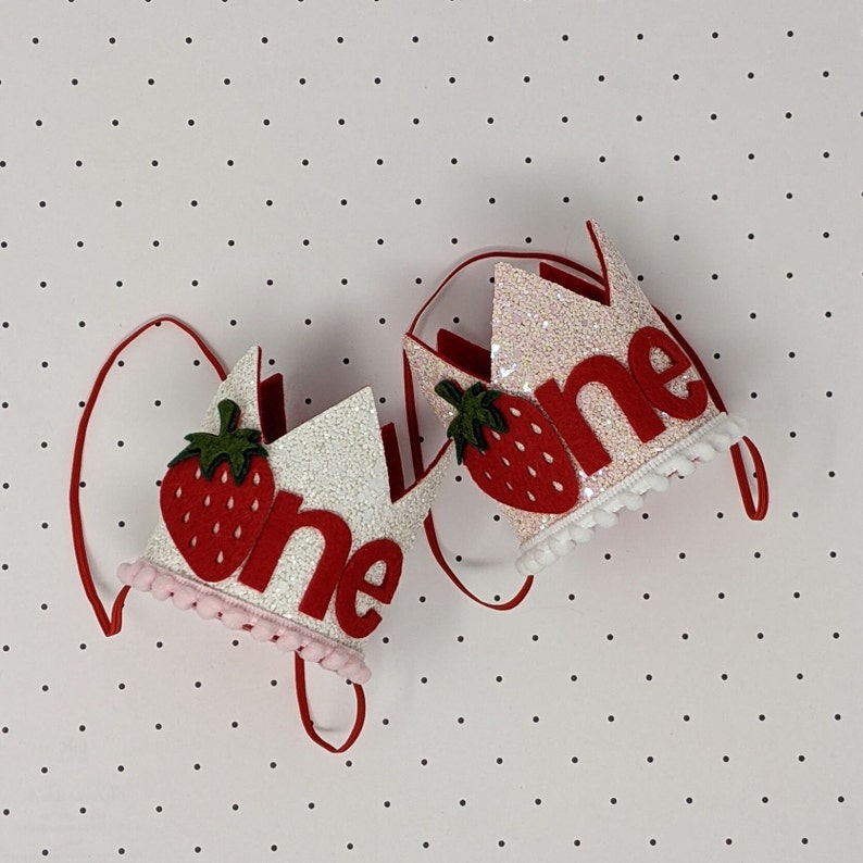May include: Two white felt crowns with red felt strawberries and the word "one" in red felt letters. The crowns have pink and white pom pom trim.
