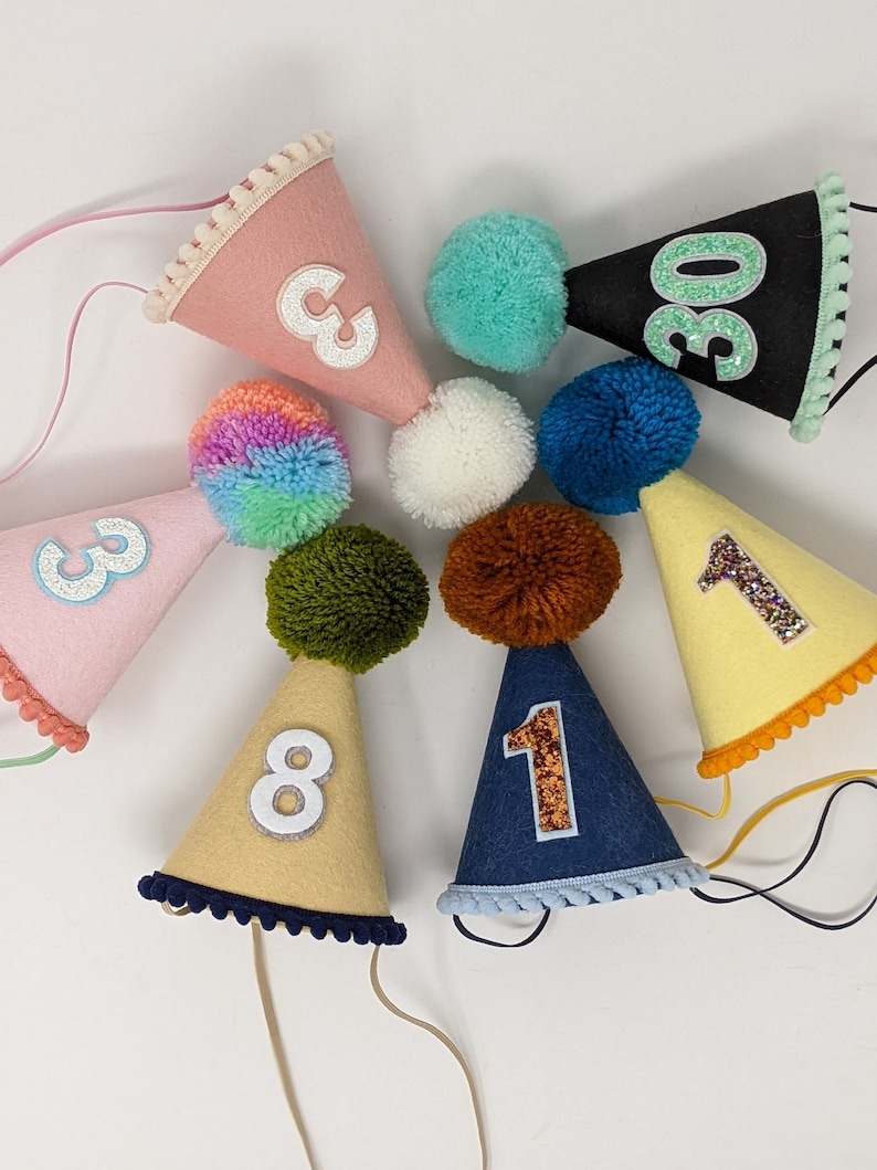 May include: A set of nine felt party hats with numbers 1, 1, 3, 3, 8, and 30. The hats are decorated with glitter and pom-poms.