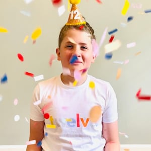 May include: A young person wearing a white t-shirt with the word "twelve" printed on it in rainbow colors. They are wearing a yellow party hat with the number "12" on it. Confetti is falling around them.