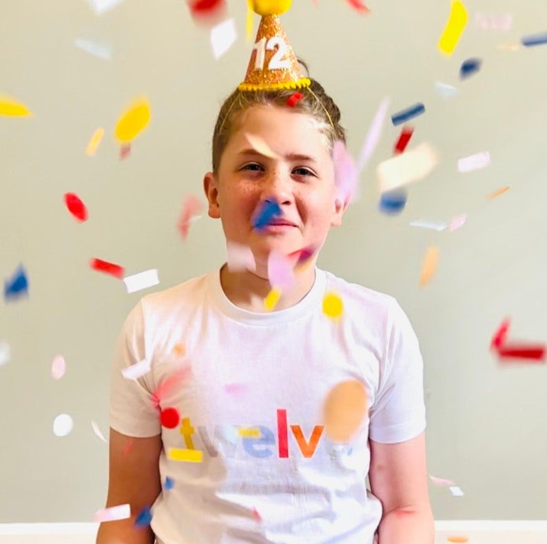 May include: A young person wearing a white t-shirt with the word "twelve" printed on it in rainbow colors. They are wearing a yellow party hat with the number "12" on it. Confetti is falling around them.