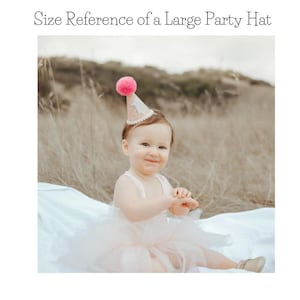 May include: A pink and gold glitter party hat with a pink pom-pom on top. The hat has a white number "1" on the front. A young child is wearing the hat.