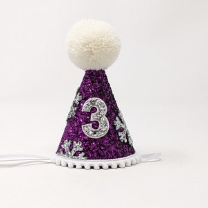 May include: Purple glitter party hat with a white pom pom on top. The hat has a silver number 3 and silver snowflake accents.