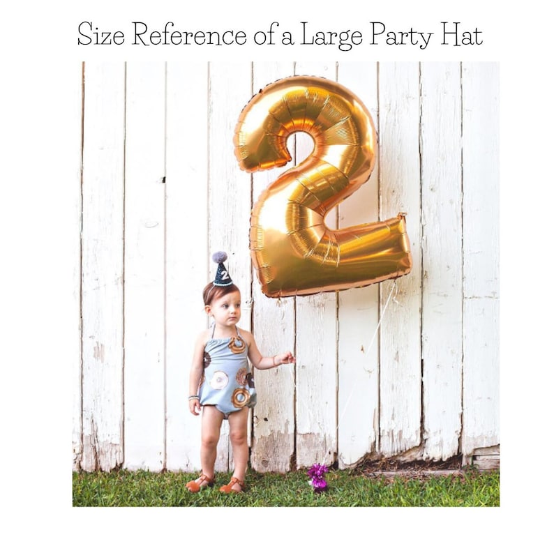 May include: A child wearing a blue and brown donut-patterned onesie and a party hat with a black pom-pom is standing in front of a white wooden fence. A large gold number 2 balloon is floating behind the child.
