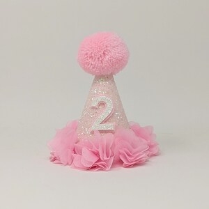 May include: Pink glitter party hat with a white number two and a pink pom pom on top. The hat has a pink tulle ruffle around the bottom.