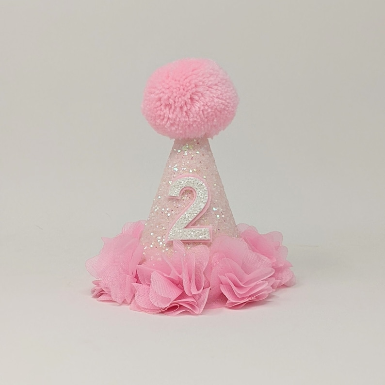 May include: Pink glitter party hat with a white number two and a pink pom pom on top. The hat has a pink tulle ruffle around the bottom.