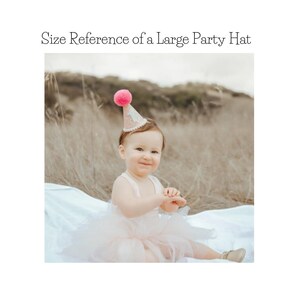 May include: A pink and white party hat with a pink pom-pom on top. The hat is worn by a young child sitting on a white blanket.