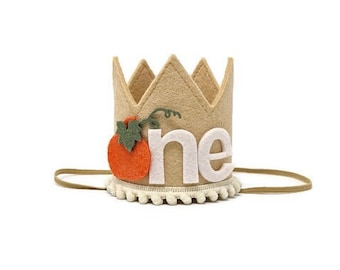 Può includere: Una corona di feltro con un "uno" in feltro bianco e una zucca arancione in feltro con un gambo verde. La corona ha un bordo di pon pon bianco e un elastico marrone.