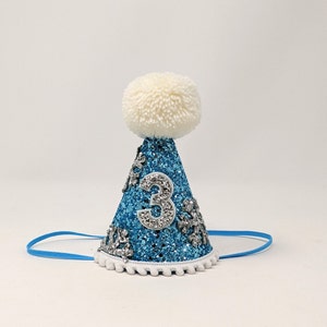 May include: A blue and silver glitter party hat with a white pom-pom on top. The hat has a white number "3" on the front and a blue elastic band.