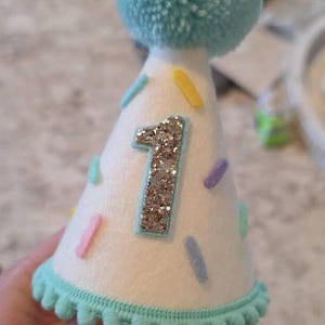 May include: A white felt party hat with a glittery silver number "1" and colourful confetti-like decorations. The hat has a light blue pom-pom on top and a matching trim around the base. A festive accessory for a first birthday celebration.