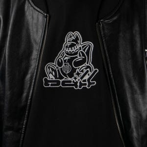 May include: Black bomber jacket with a black hoodie underneath. The hoodie features a white embroidered graphic of a cartoon character and the text "SC24". The jacket has a zipper closure.