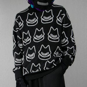 May include: Black sweater with a repeating white cat face pattern. The sweater has a black turtleneck and cuffs. The cat face design features a smiling mouth and a stylized cat head. The sweater is worn with black pants and a patterned neck covering.