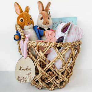 May include: A wicker basket filled with a pink and white blanket, sunglasses, a pink bow, and two plush bunny toys. A wooden tag with the name "Kendra" is attached to the basket.