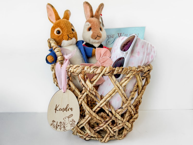 May include: A wicker basket filled with a pink and white blanket, sunglasses, a pink bow, and two plush bunny toys. A wooden tag with the name "Kendra" is attached to the basket.