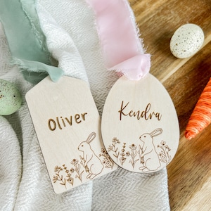 May include: Two wooden Easter egg-shaped place cards with bunny illustrations and floral designs. The cards are personalised with the names "Oliver" and "Kendra".