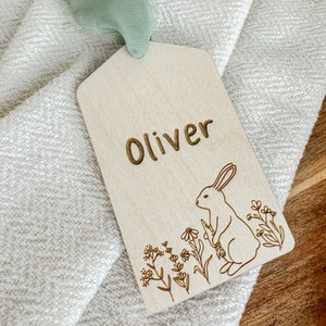 May include: A wooden tag with the name "Oliver" engraved on it. The tag features a small rabbit illustration with flowers and greenery.