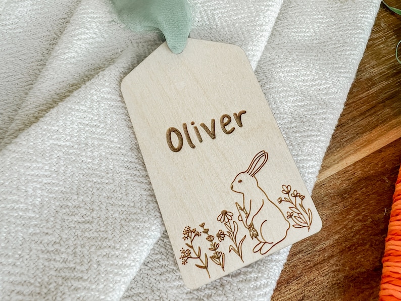May include: A wooden tag with the name "Oliver" engraved on it. The tag features a small rabbit illustration with flowers and greenery.