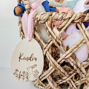 May include: A wooden Easter egg ornament with the name Kendra engraved on it. The egg is decorated with a small rabbit and flowers. The egg is hanging from a pink ribbon on a wicker basket filled with soft toys and other Easter decorations.