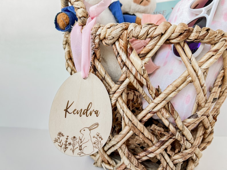 May include: A wooden Easter egg ornament with the name Kendra engraved on it. The egg is decorated with a small rabbit and flowers. The egg is hanging from a pink ribbon on a wicker basket filled with soft toys and other Easter decorations.
