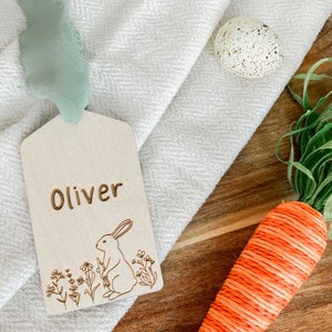 May include: A wooden tag with the name "Oliver" engraved on it. The tag features a laser-cut design of a rabbit sitting in a field of flowers.
