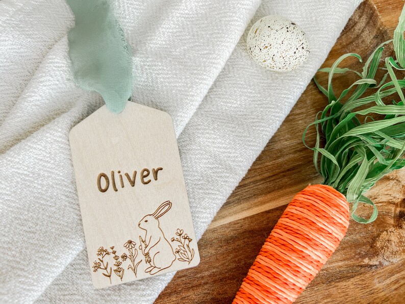 May include: A wooden tag with the name "Oliver" engraved on it. The tag features a laser-cut design of a rabbit sitting in a field of flowers.