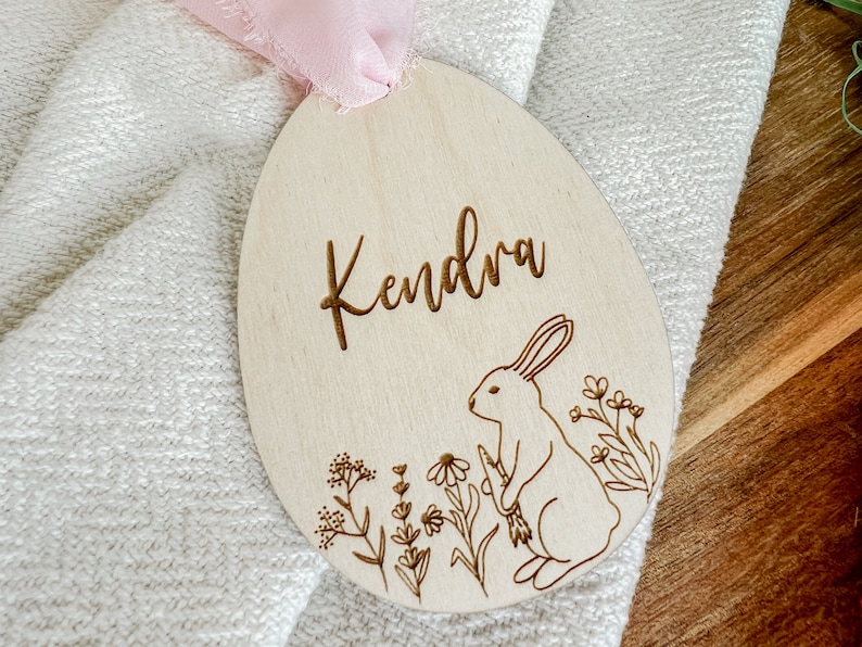 May include: A wooden Easter egg ornament with the name "Kendra" engraved on it. The ornament features a bunny rabbit and floral designs.