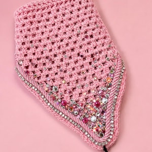 May include: A pink crocheted accessory, trimmed with a decorative edge and adorned with rhinestones and beads. It has a black string attached. The background is a solid pink colour.