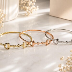 May include: Three metal bangle bracelets featuring a horse bit design. The set includes one gold-tone, one rose gold-tone, and one silver-tone bracelet, each with a polished finish and a central equestrian-inspired link detail.