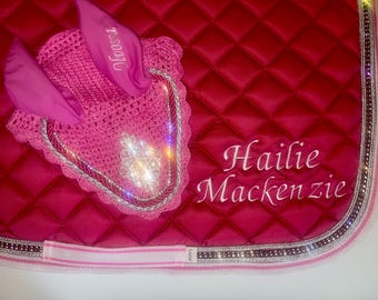 May include: A quilted, hot pink saddle pad with a matching pink crochet ear bonnet. The ear bonnet has the name "Noosa" embroidered on it. The saddle pad is embroidered with the name "Hailie Mackenzie".